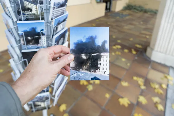 A hand holding a postcard in front of a postcard stand, in front of a floor full of autumn leaves. The postcard depicts a big old building with a lot of black smoke