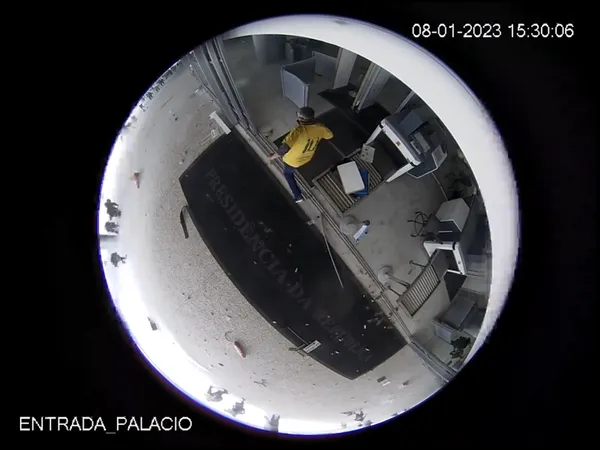 A film still: A fischeye view (a rounded image over a black background), probably from a security camera, with a person with a yellow jacked walking in a vandalized hall. On the bottom, the text Entrada Palacio
